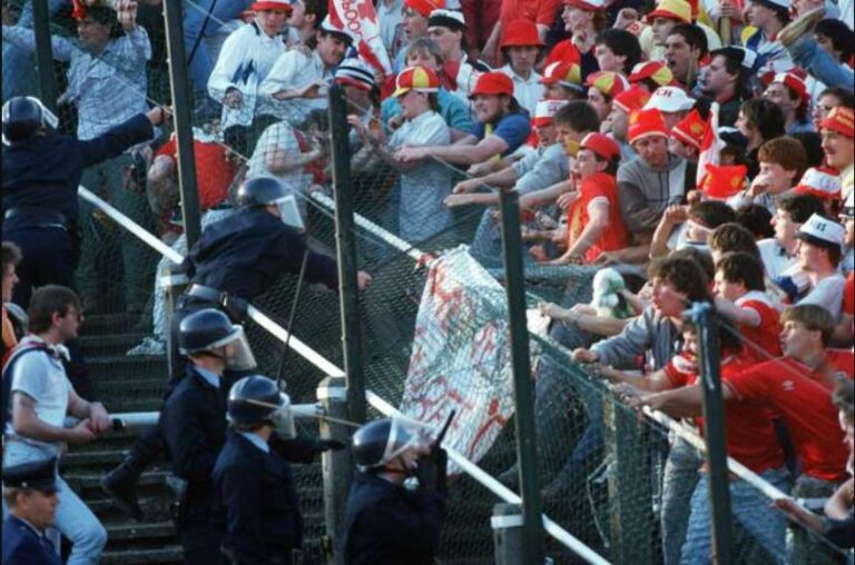 An image of a football stadium disaster, showing football fans beginning to riot behind a metal chain-link fence, with riot police pushing back against them.