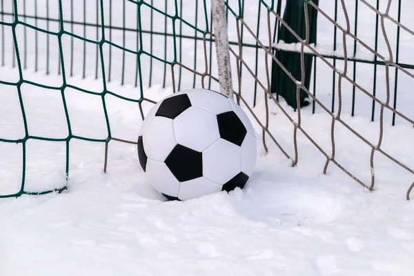 Winter Football: An image with more of a close up frame, showing a football sitting on a mound of snow, placed in the corner of a goal with dark green netting